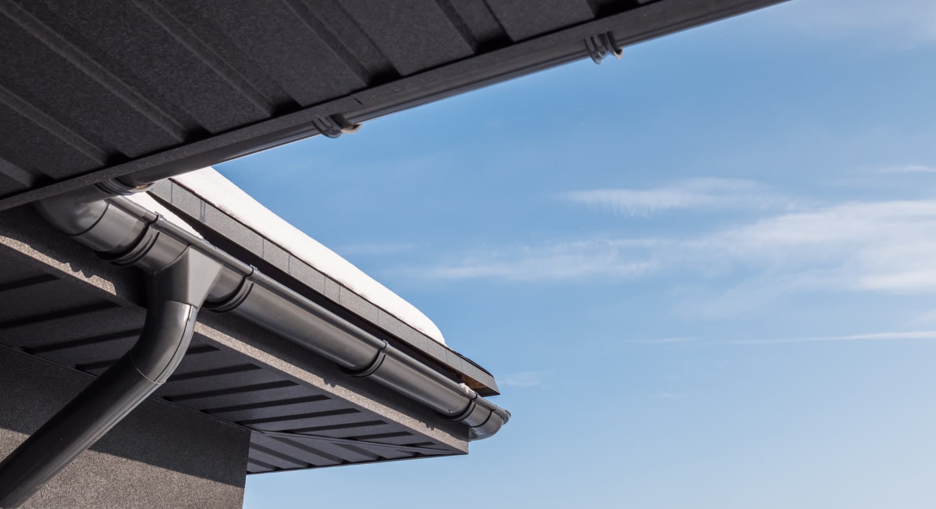 Dark seamless guttering and downpipe running along a roof edge against a blue sky